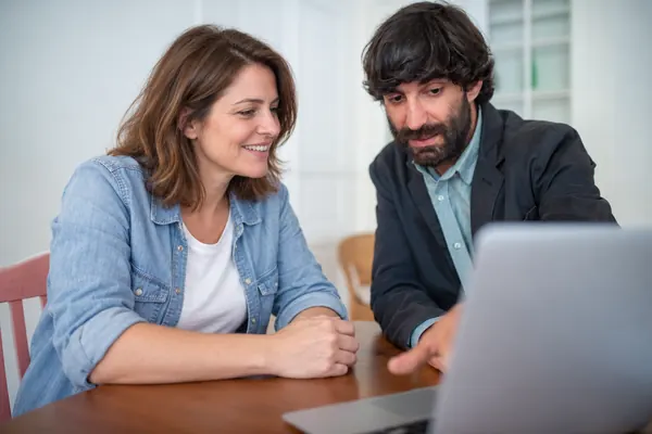 A man shows something on a laptop to a woman in a denim shirt, who smiles seated at a wooden table.