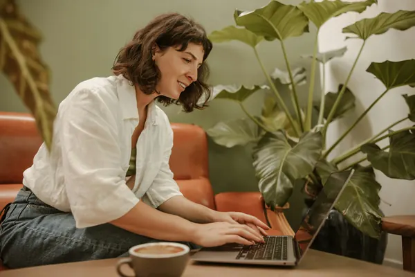 Una mujer sonríe mientras trabaja con su portátil en un sofá, junto a una planta.