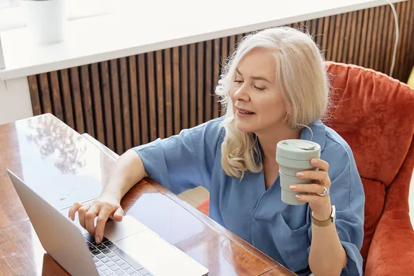A woman with a mug in hand works on her laptop by the window.