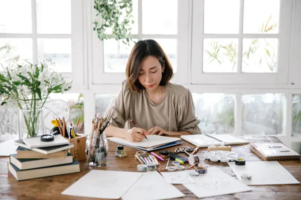 A woman draws in a notebook, at a table covered in papers, pencils and books.