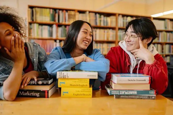 Three students laugh together in a library, with German dictionaries on the table.